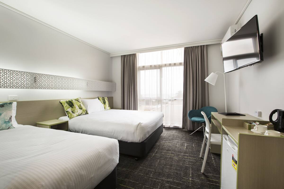 A hotel room with two beds, a desk, a chair, a TV, and a window with curtains.
