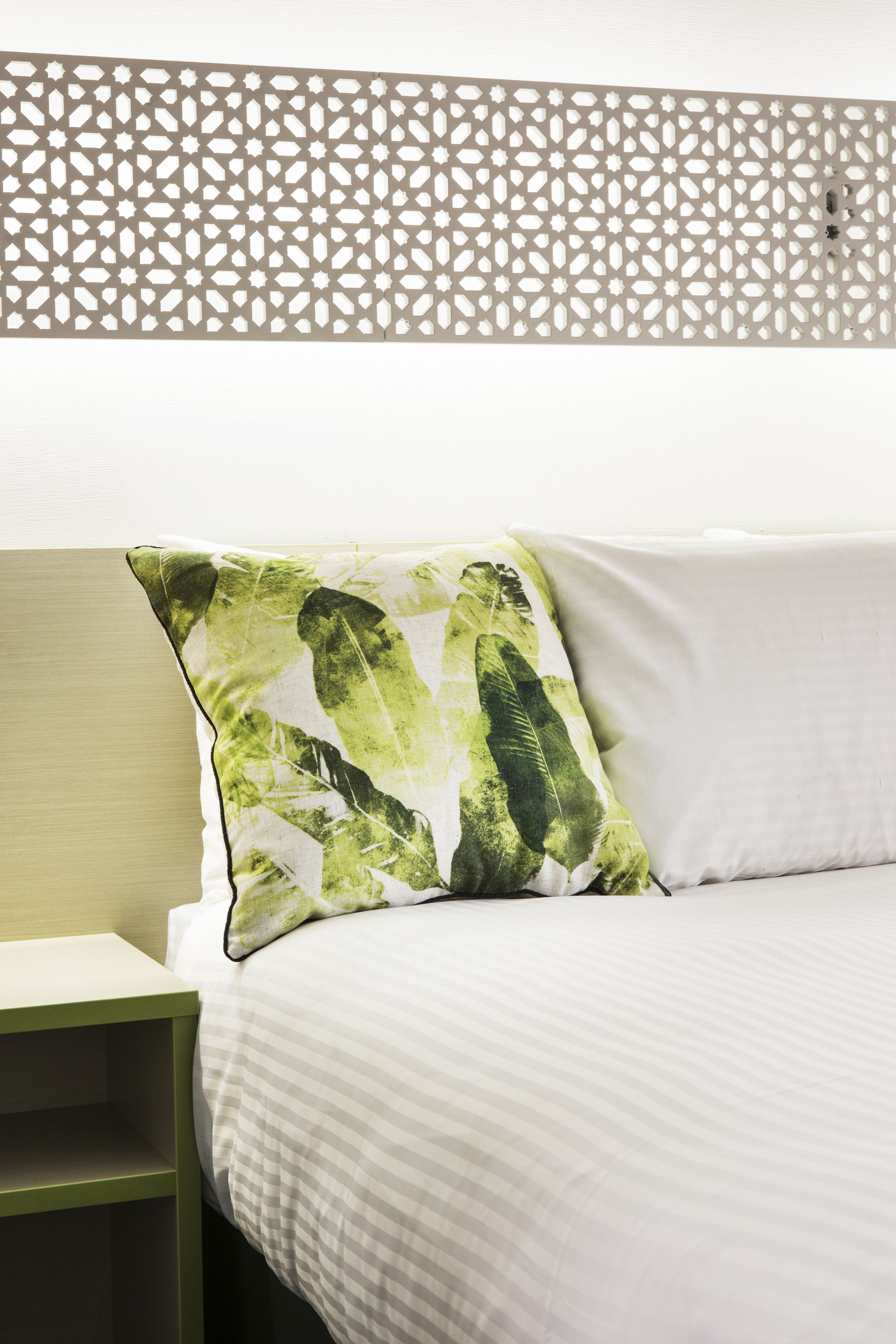 A bed with a white pillow and a green leaf-patterned pillow, a wooden headboard, and a light-colored wall.