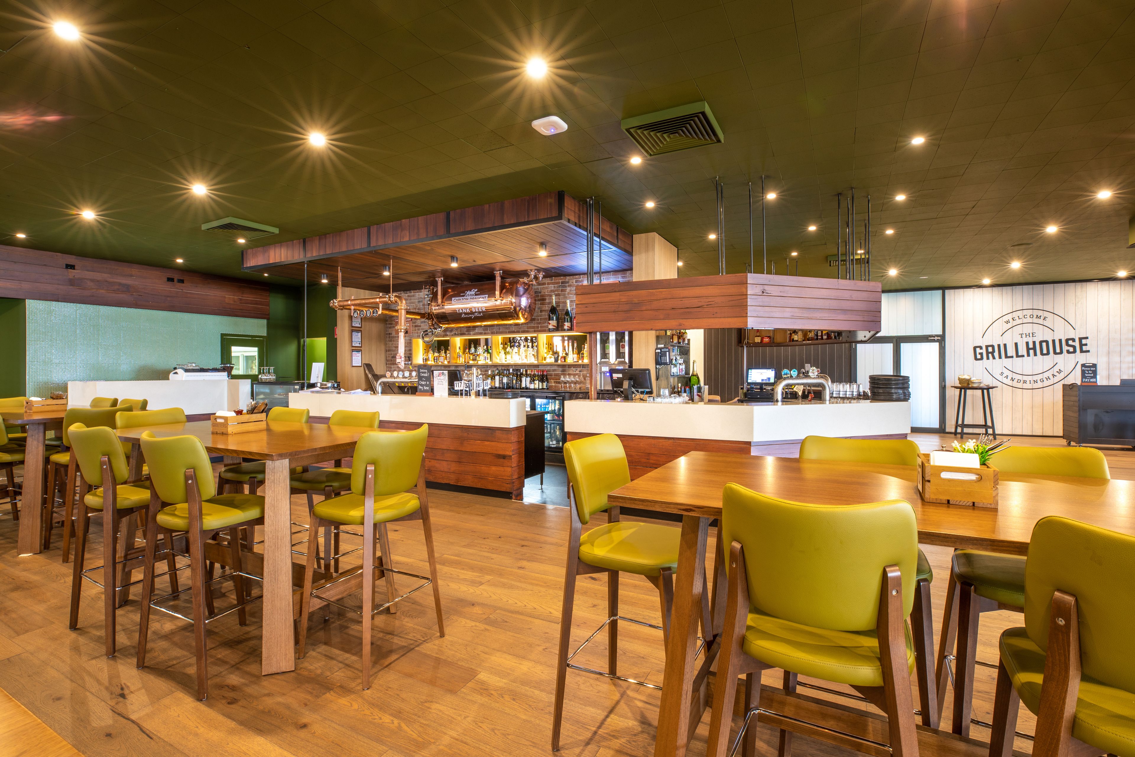 Un restaurante con mesas de madera y sillas verdes, una zona de bar y un letrero que dice 'GRILLHOUSE'.