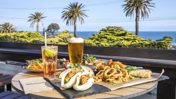 A table with food and drinks, palm trees, and a view of the ocean. - Nightcap at Sandringham Hotel (Sandringham)