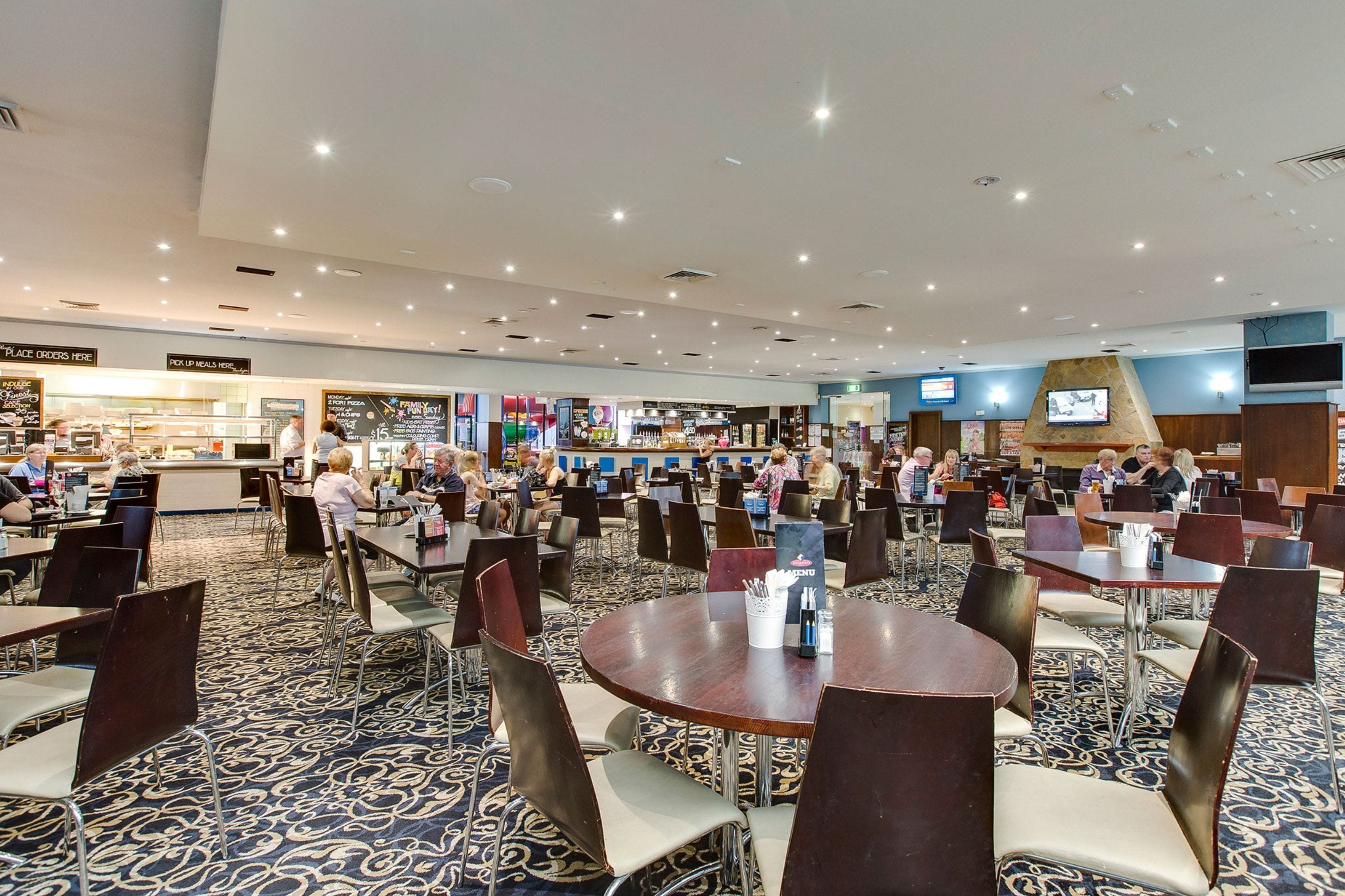 A spacious restaurant with multiple dining tables and chairs, a counter for ordering, and a television screen on the wall.