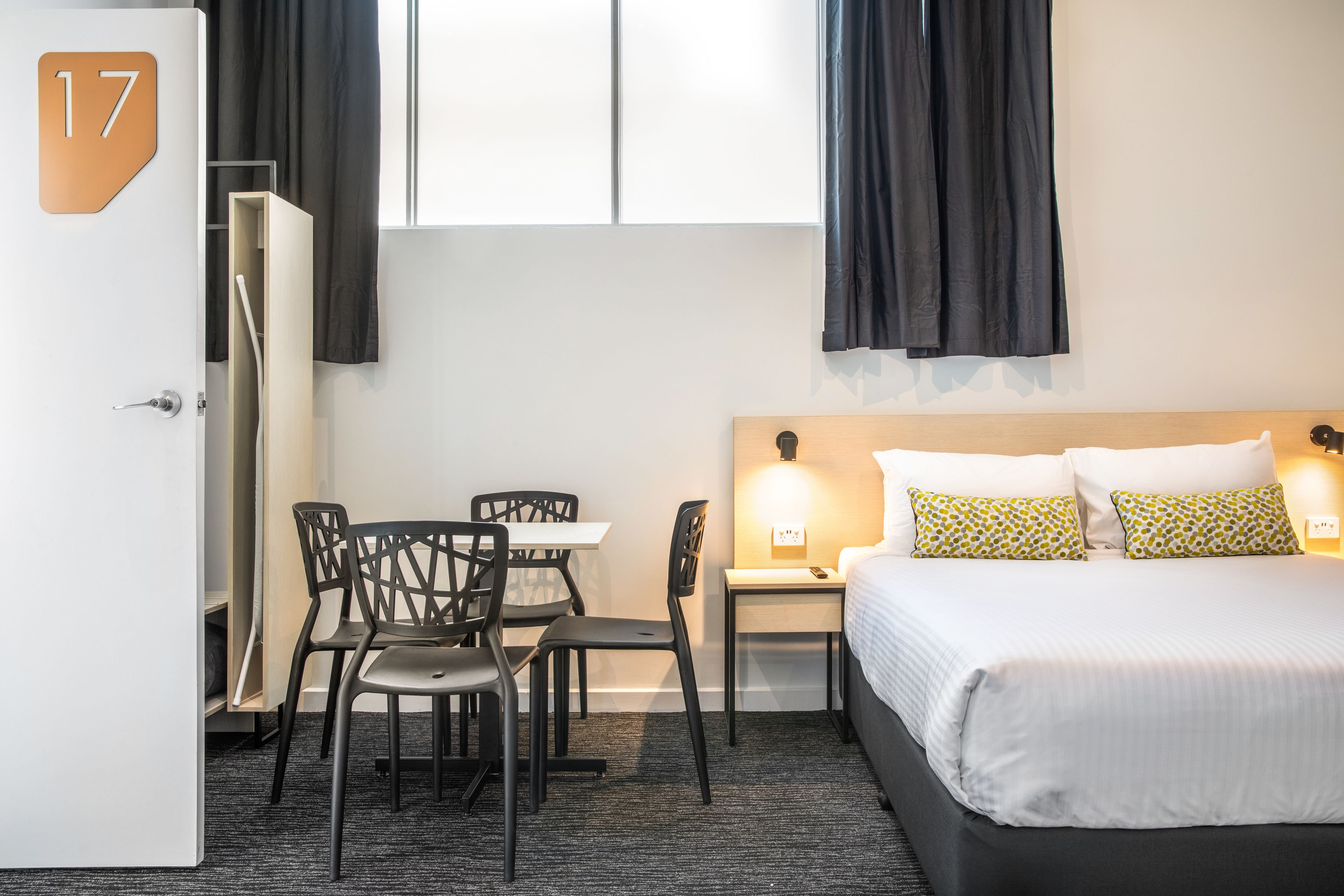 A hotel room with a bed, a desk, two chairs, a nightstand, and a window with curtains.