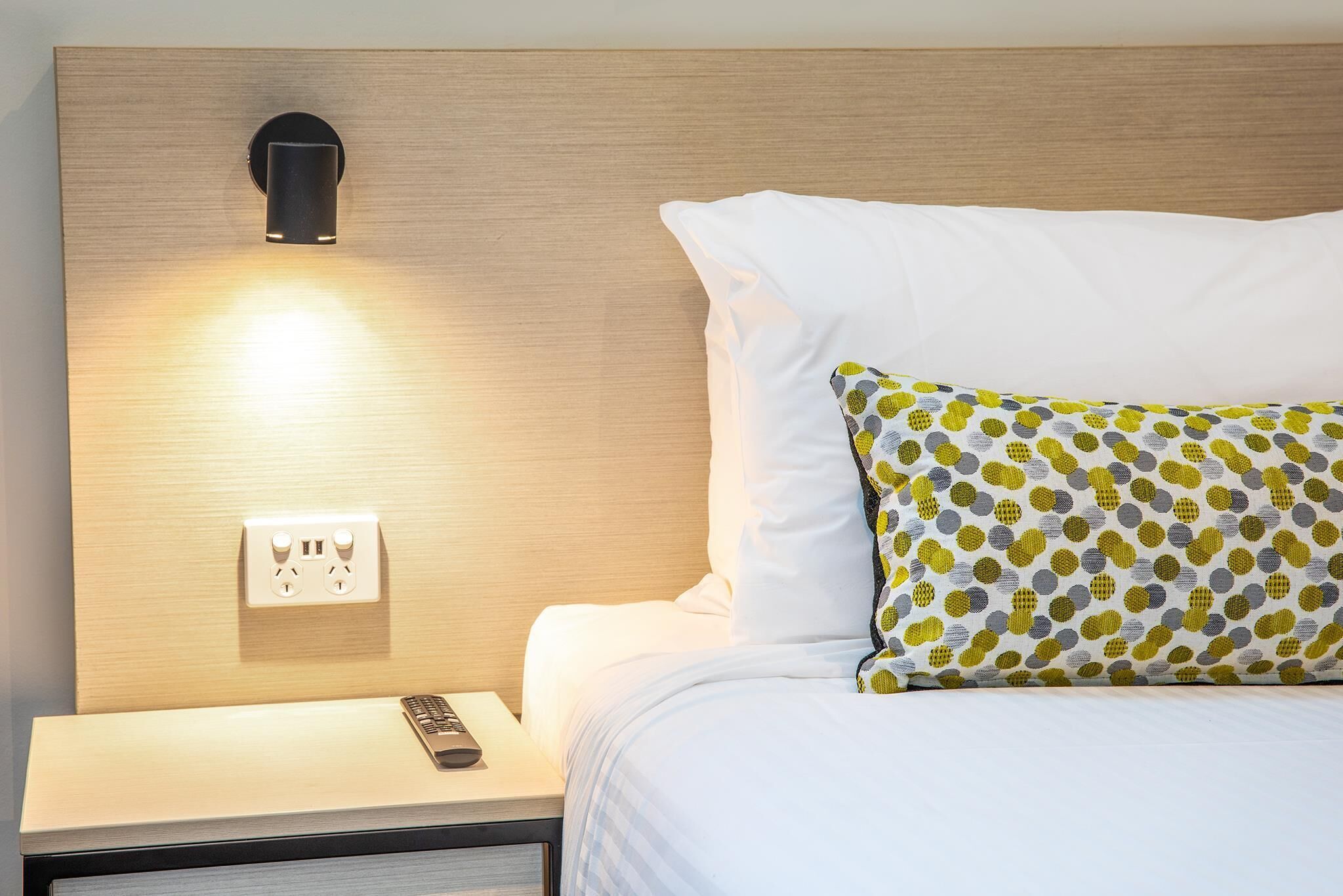 A neatly made bed with white bedding and a patterned pillow, a bedside table with a remote control, and a wall-mounted light fixture.