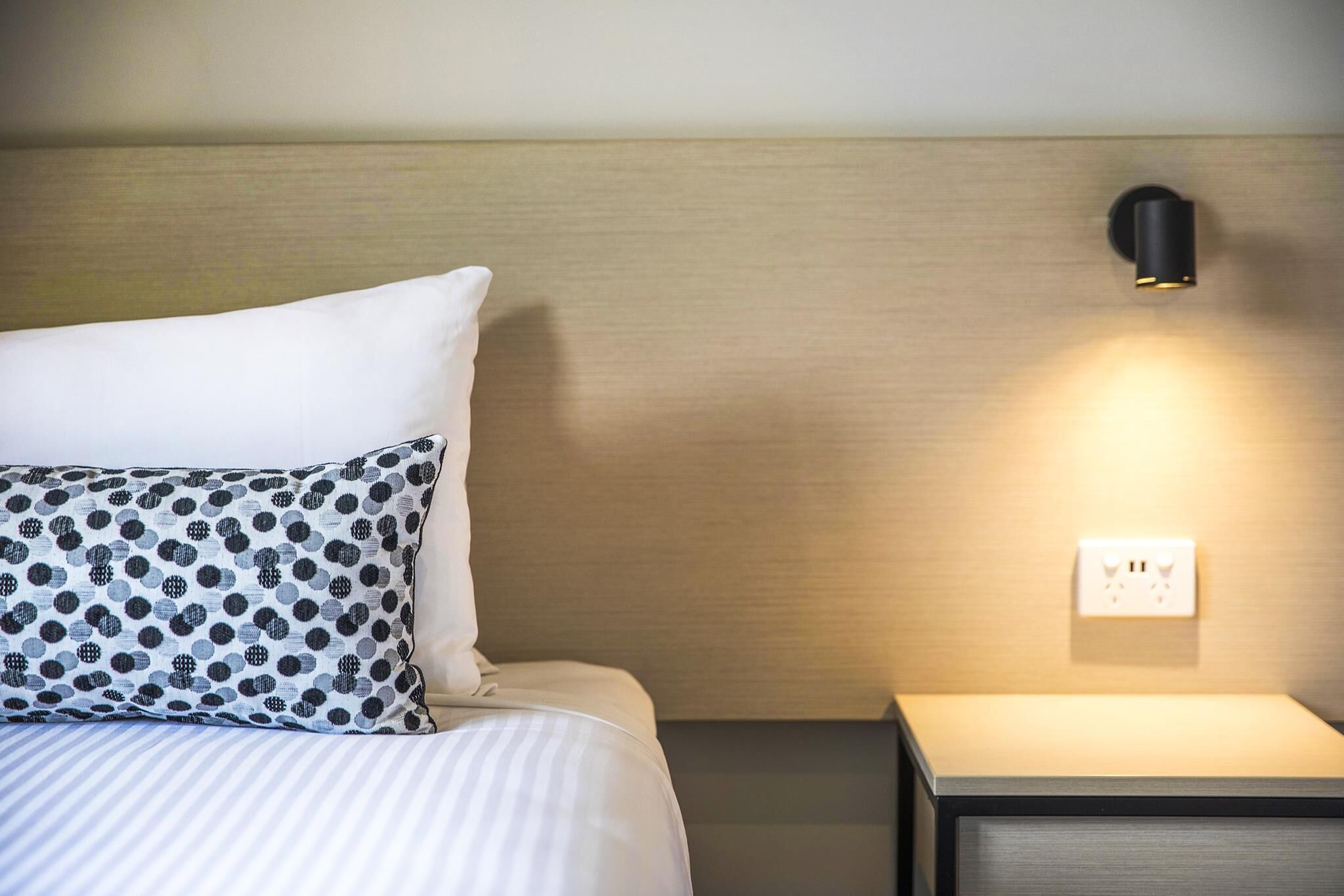 A neatly made bed with a striped bedspread and patterned pillows, a wall-mounted light, and a power outlet on a bedside table.