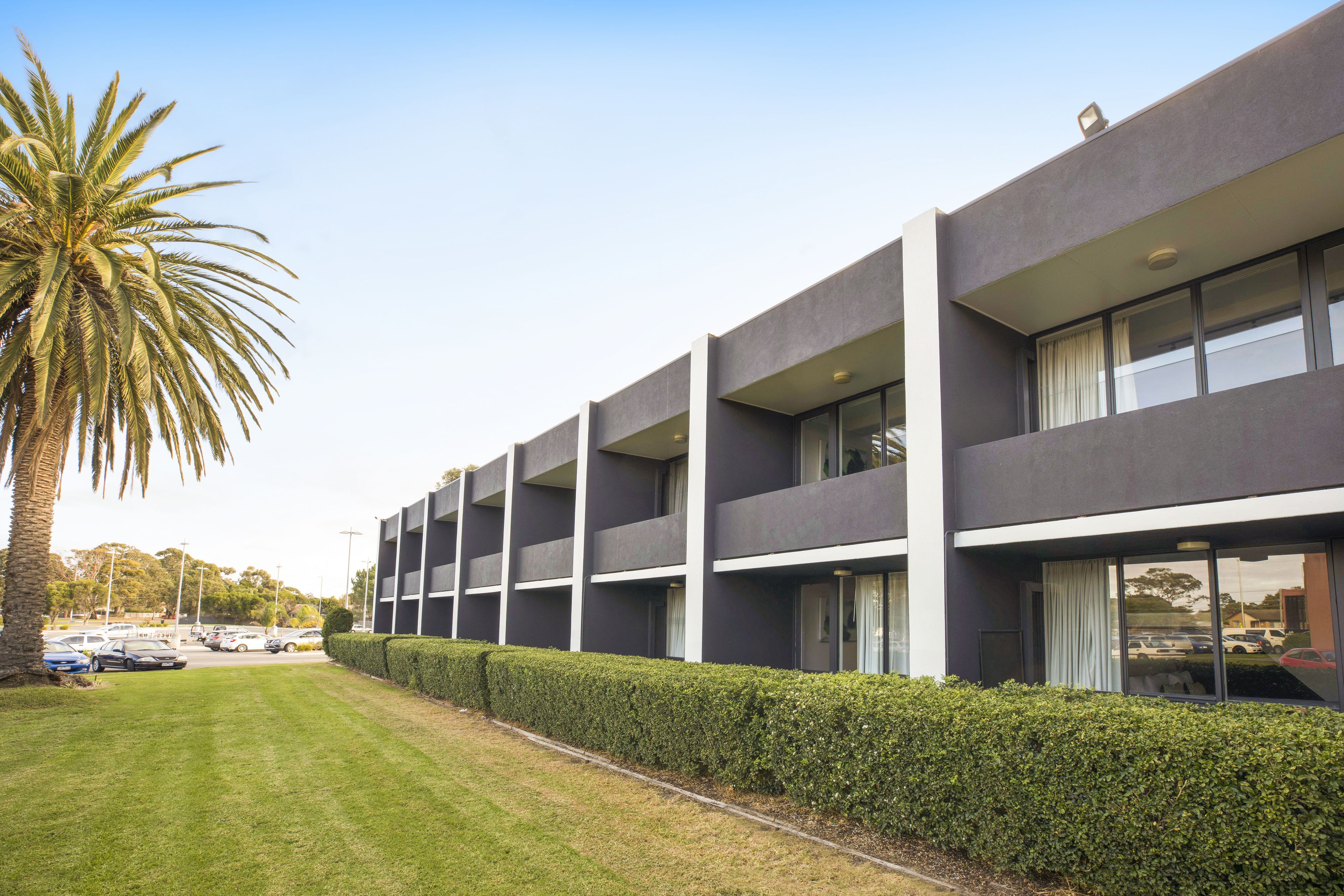 A modern multi-story building with balconies, a well-maintained lawn, and parked cars in the background.