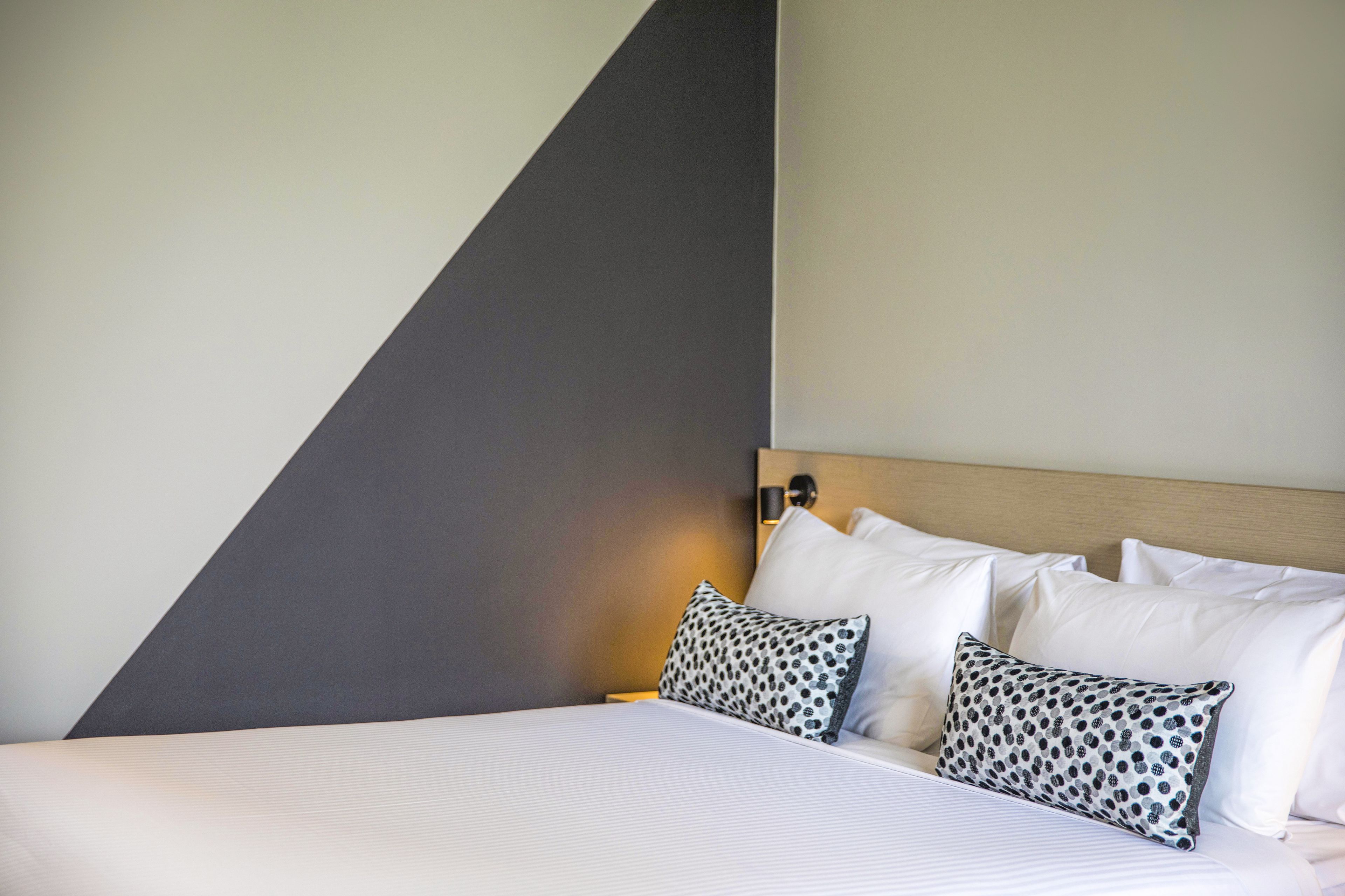 A neatly made bed with white linens and two patterned pillows, a wooden headboard, and a wall with a geometric design.