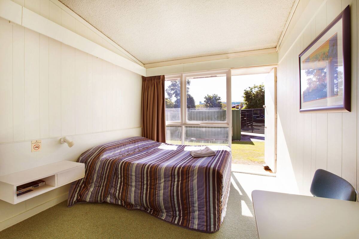 A bedroom with a bed, a desk, a window, and a door leading to an outdoor area.