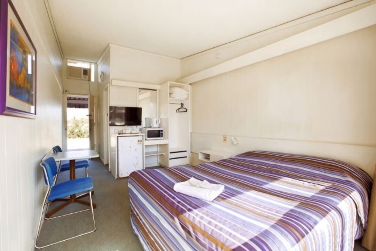 Una habitación pequeña con cama, escritorio, silla, microondas y televisor.