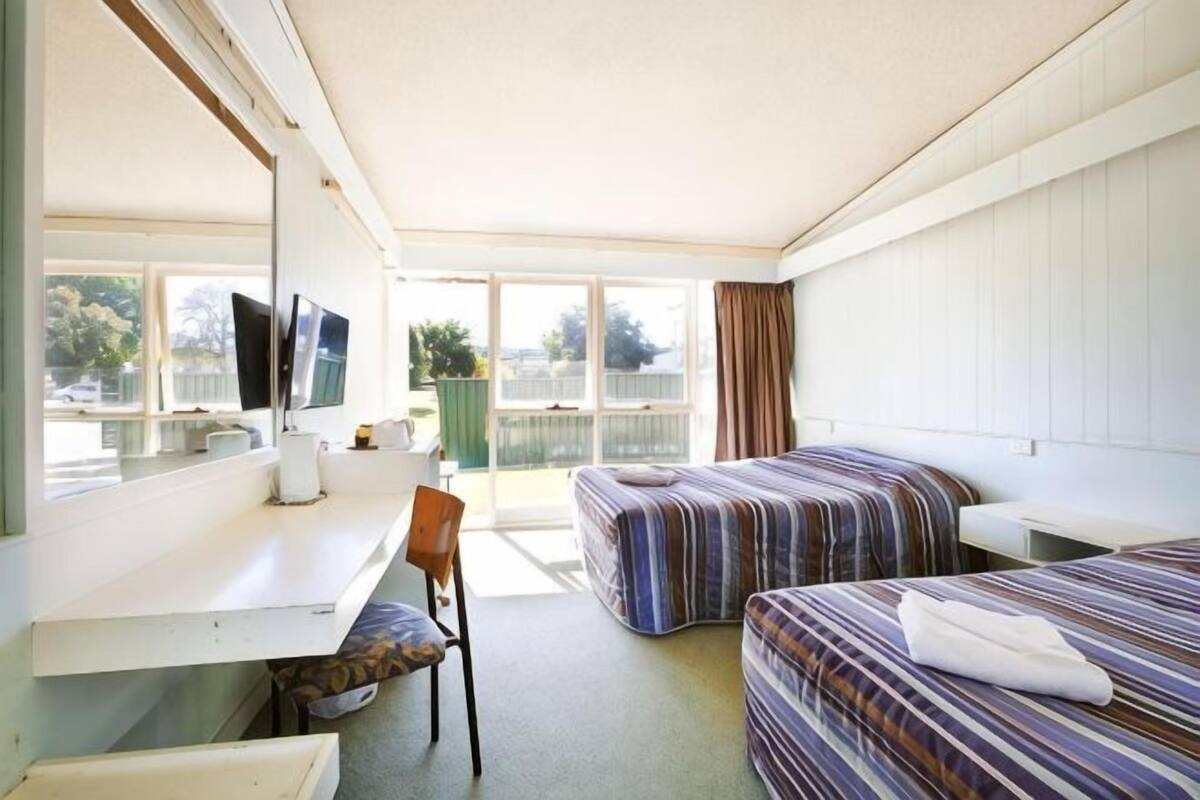 A hotel room with two beds, a desk, a chair, a TV, and large windows.