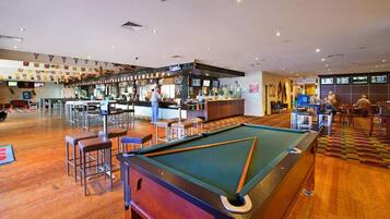 A pool table with surrounding stools, a bar area with a bartender, and a seating area with tables and chairs.