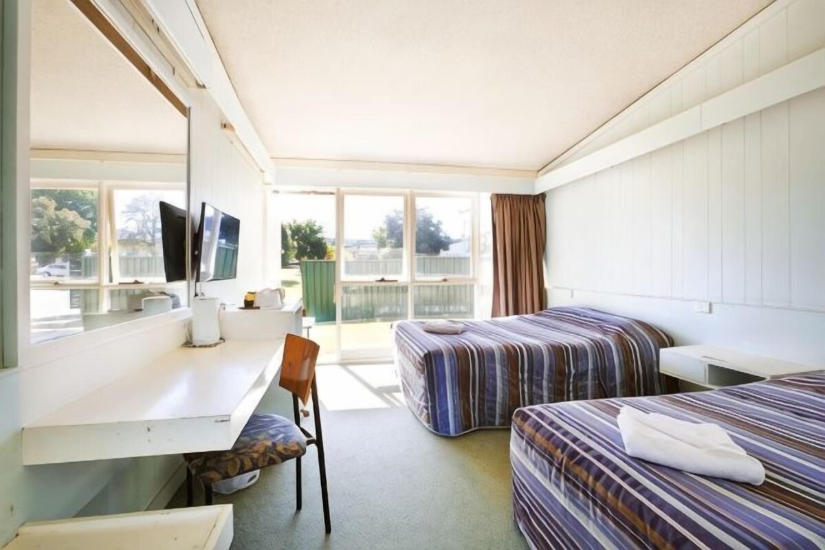 A hotel room with two beds, a desk, a chair, a TV, and large windows.