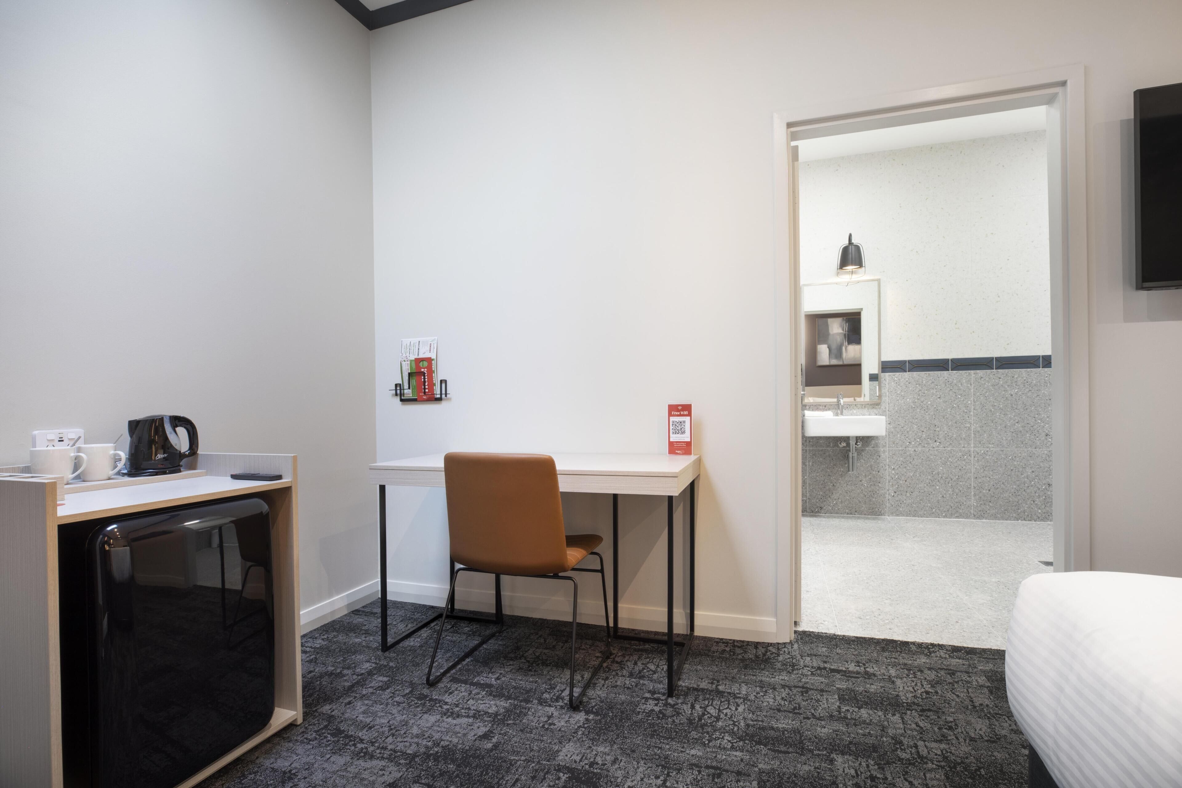 A hotel room with a desk, chair, bed, and a small kitchenette area.