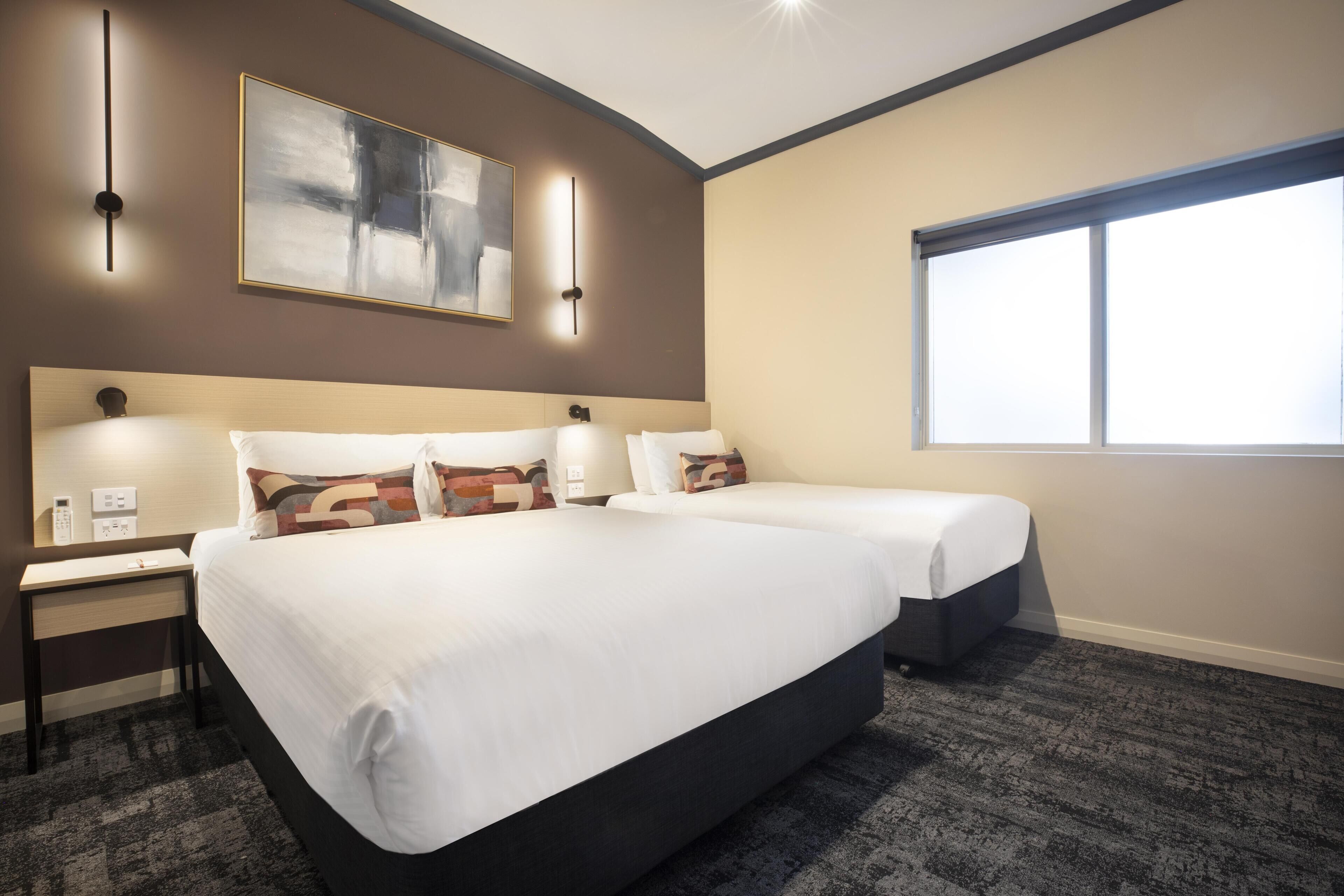 A hotel room with two beds, a desk, a chair, a large window, and a painting on the wall.