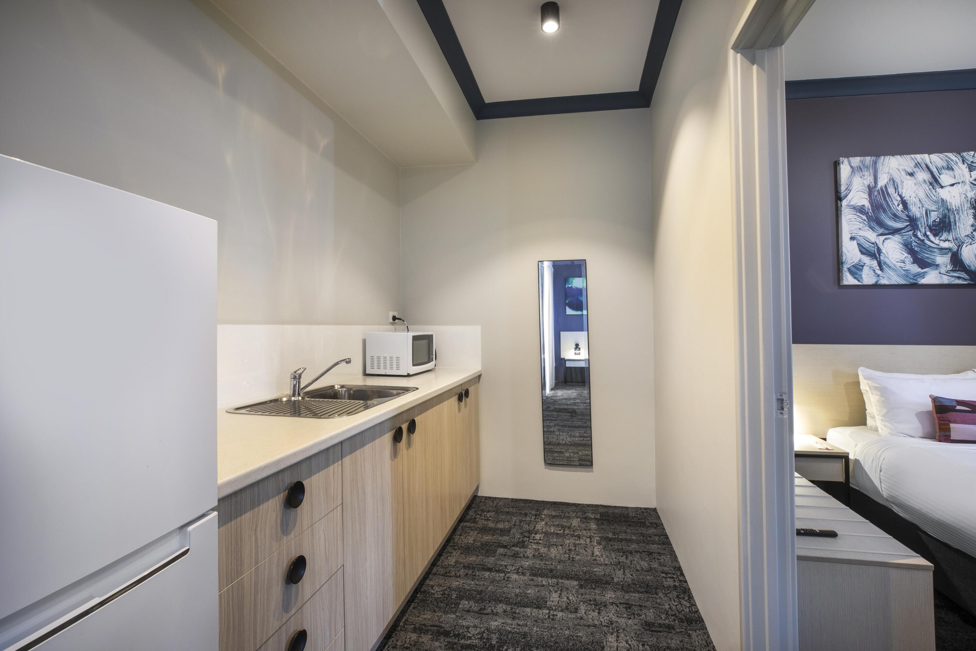 A compact hotel room with a kitchenette, a bed, a bedside table, and a mirror.