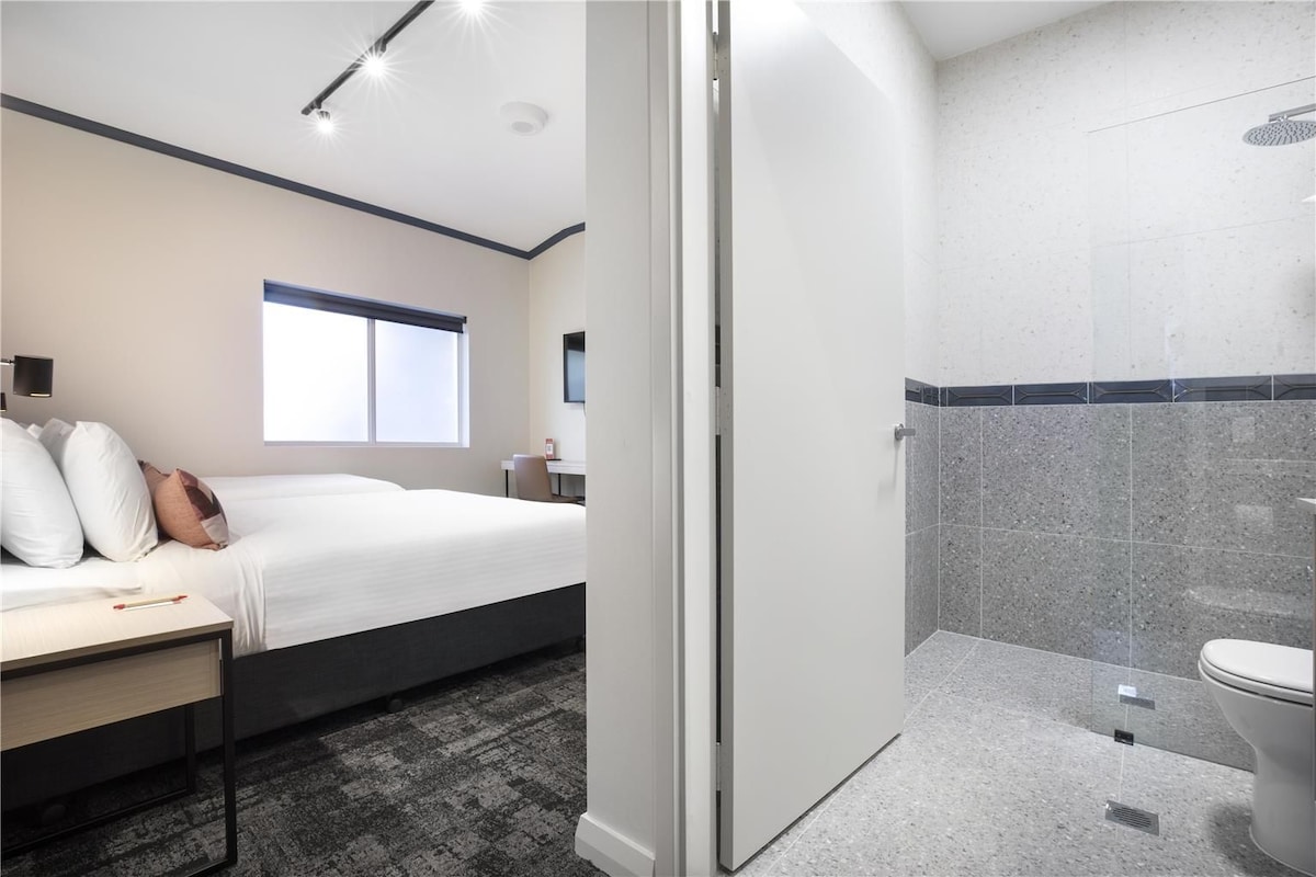 A hotel room with a bed, bedside table, and a shower area with a toilet.