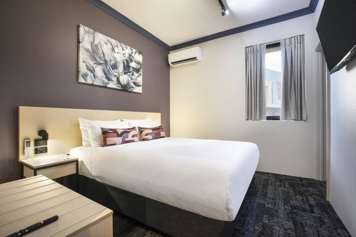 A hotel room with a bed, bedside table, nightstand, and an air conditioning unit.