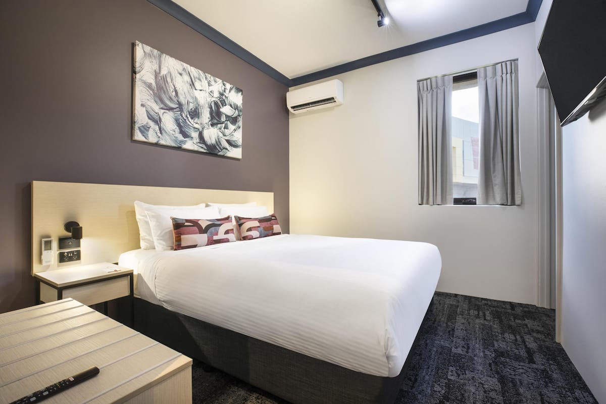 A hotel room with a bed, bedside table, nightstand, and an air conditioning unit.