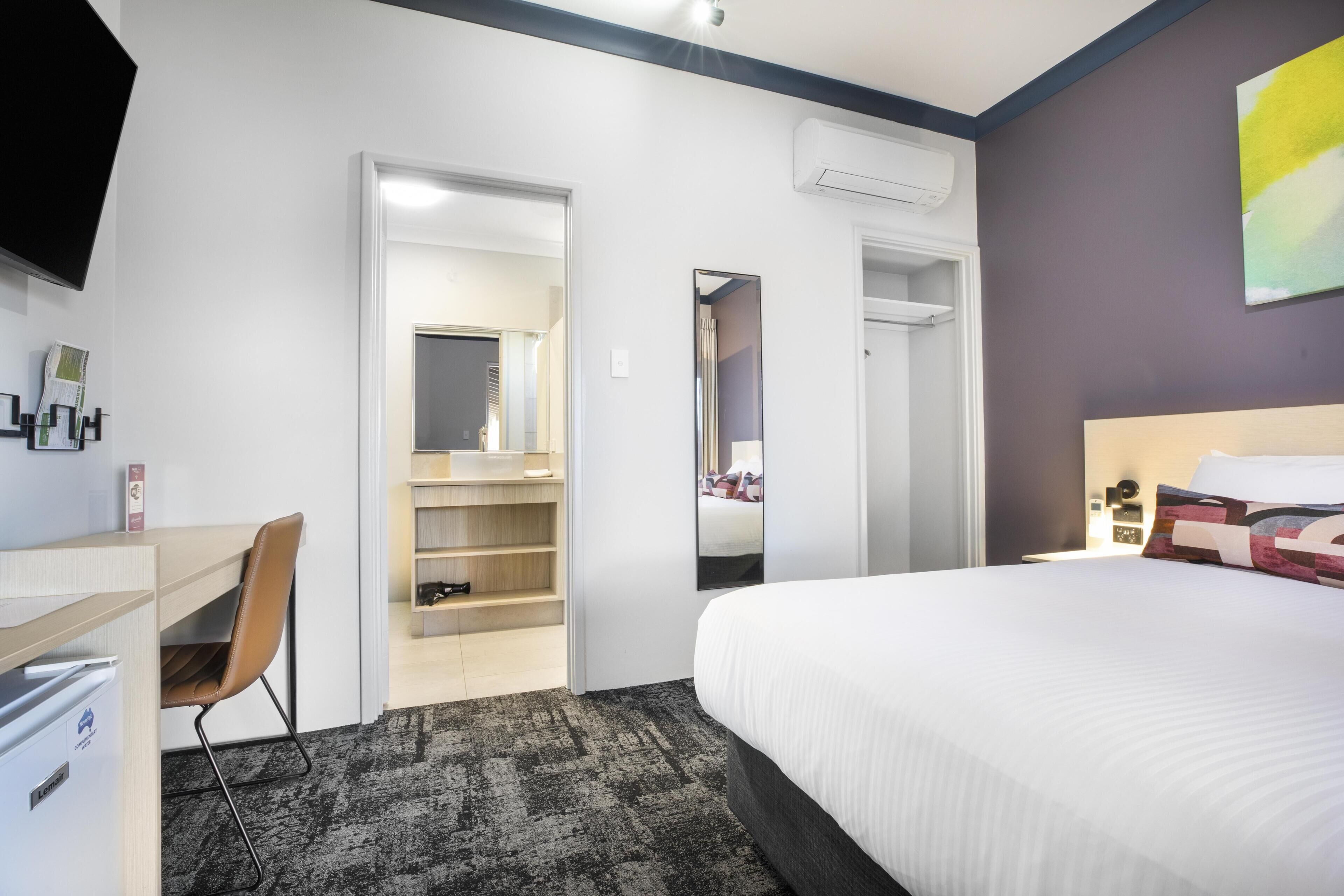 A hotel room with a bed, a desk, a chair, a wardrobe, and a mirror.