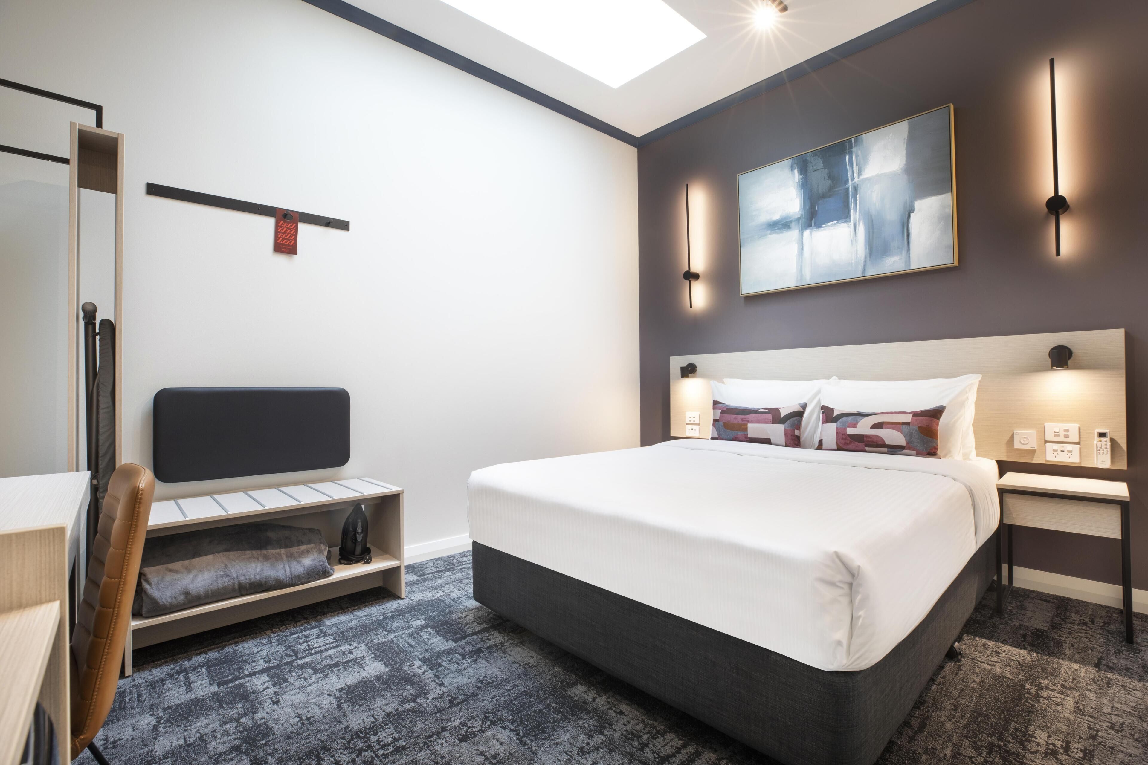 A modern hotel room with a bed, a desk, a chair, a shelf, and a painting on the wall.