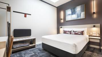 A modern hotel room with a bed, a desk, a chair, a shelf, and a painting on the wall.
