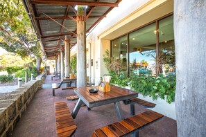 An outdoor patio with wooden tables and benches, surrounded by greenery and a covered walkway.