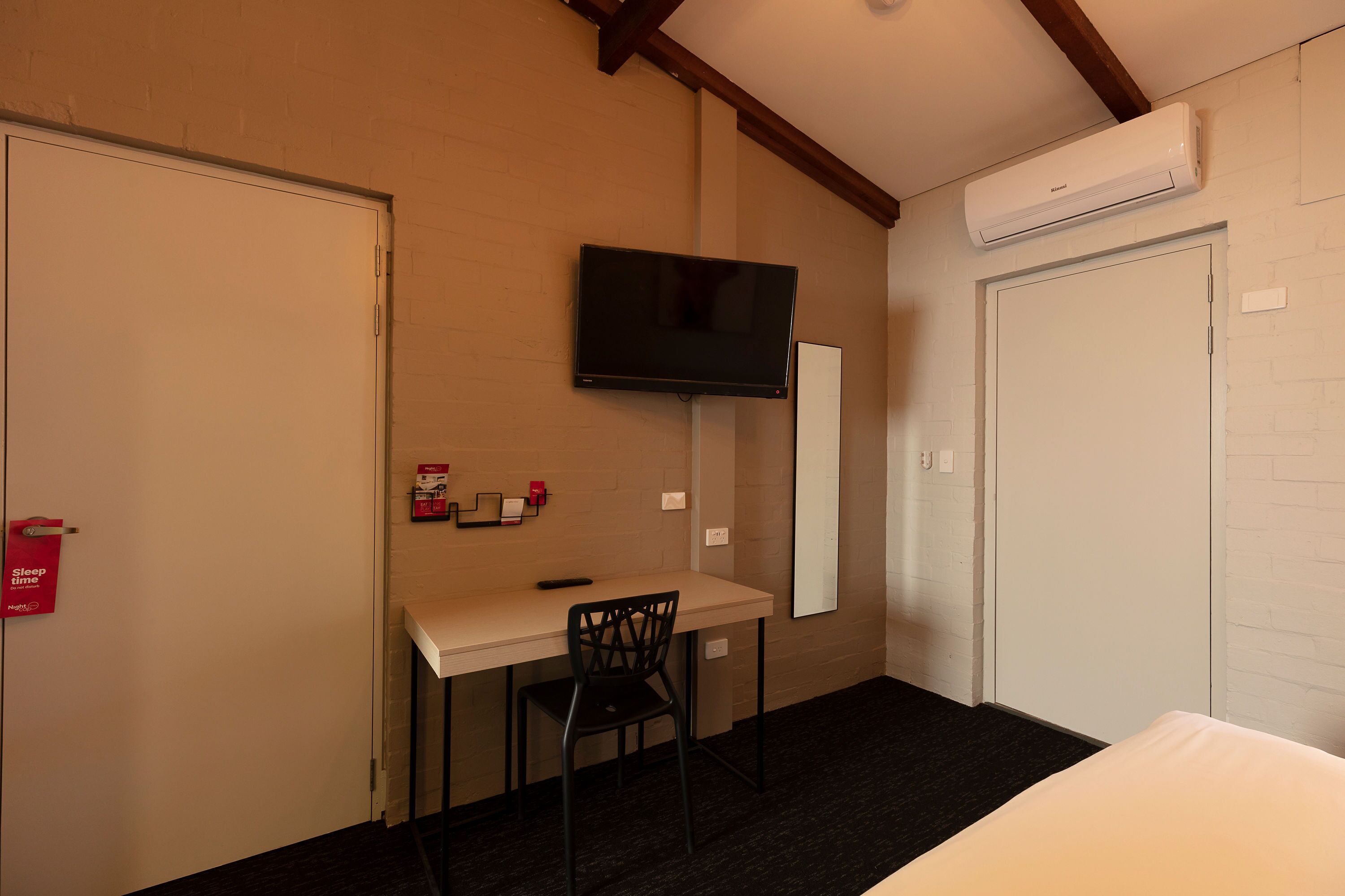 A hotel room with a bed, a desk with a chair, a television mounted on the wall, and an air conditioning unit.