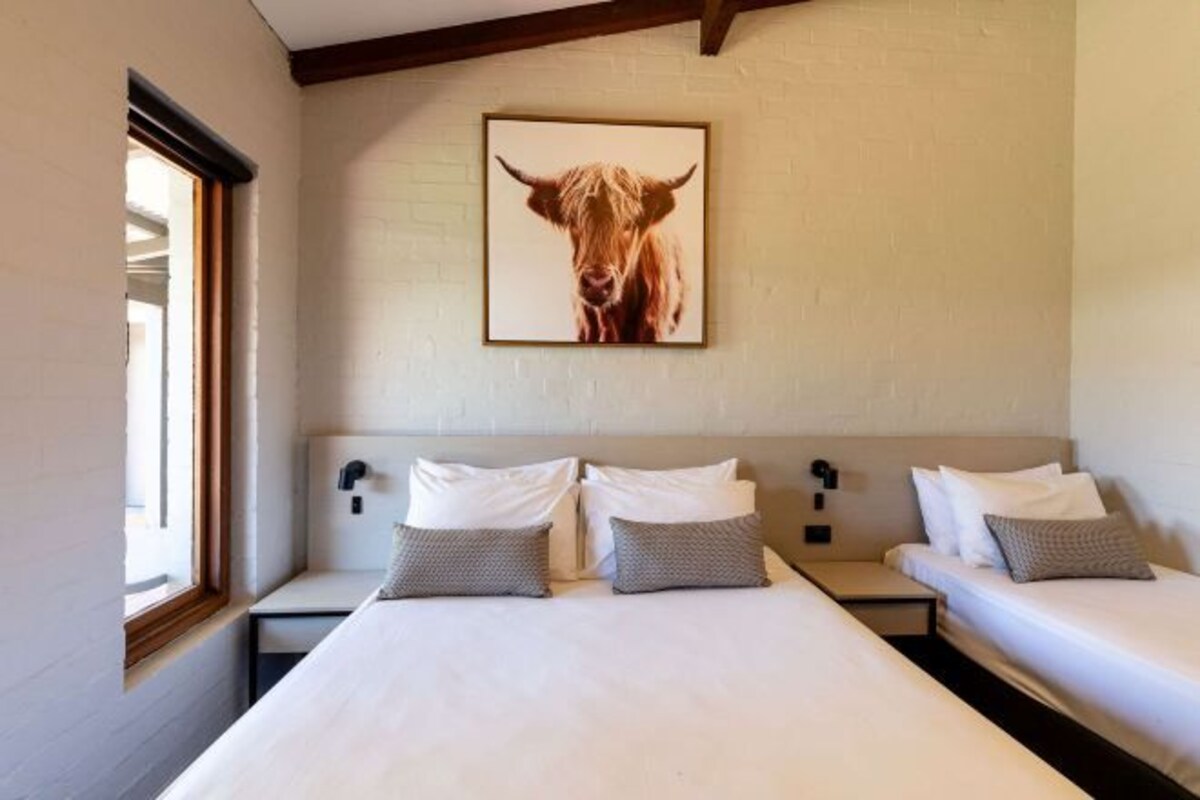 A hotel room with two beds, a large framed picture of a yak on the wall, and a window on the left side.