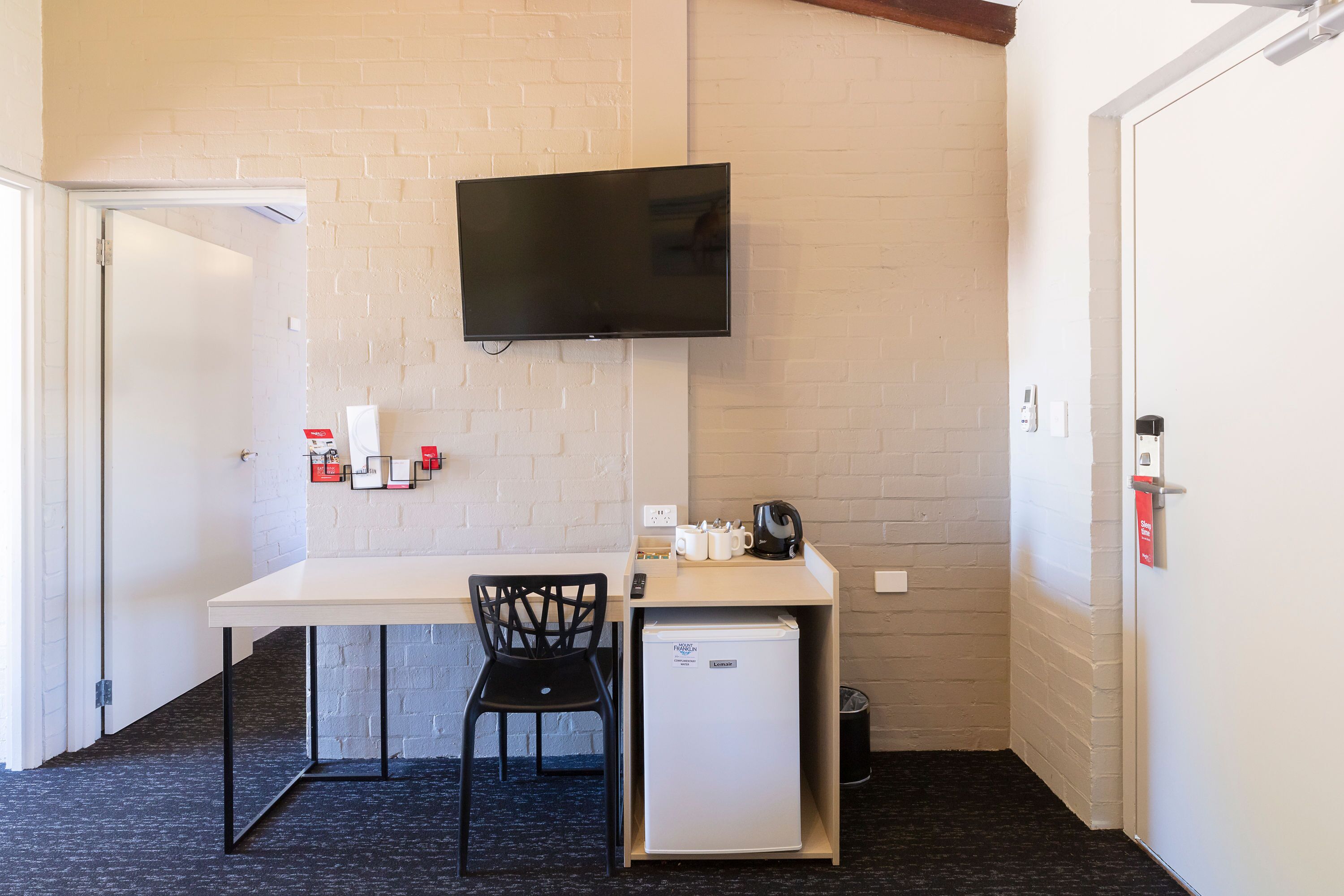 A compact room with a white brick wall, a flat-screen TV mounted above a desk, a small refrigerator, a black chair, and a coffee machine.