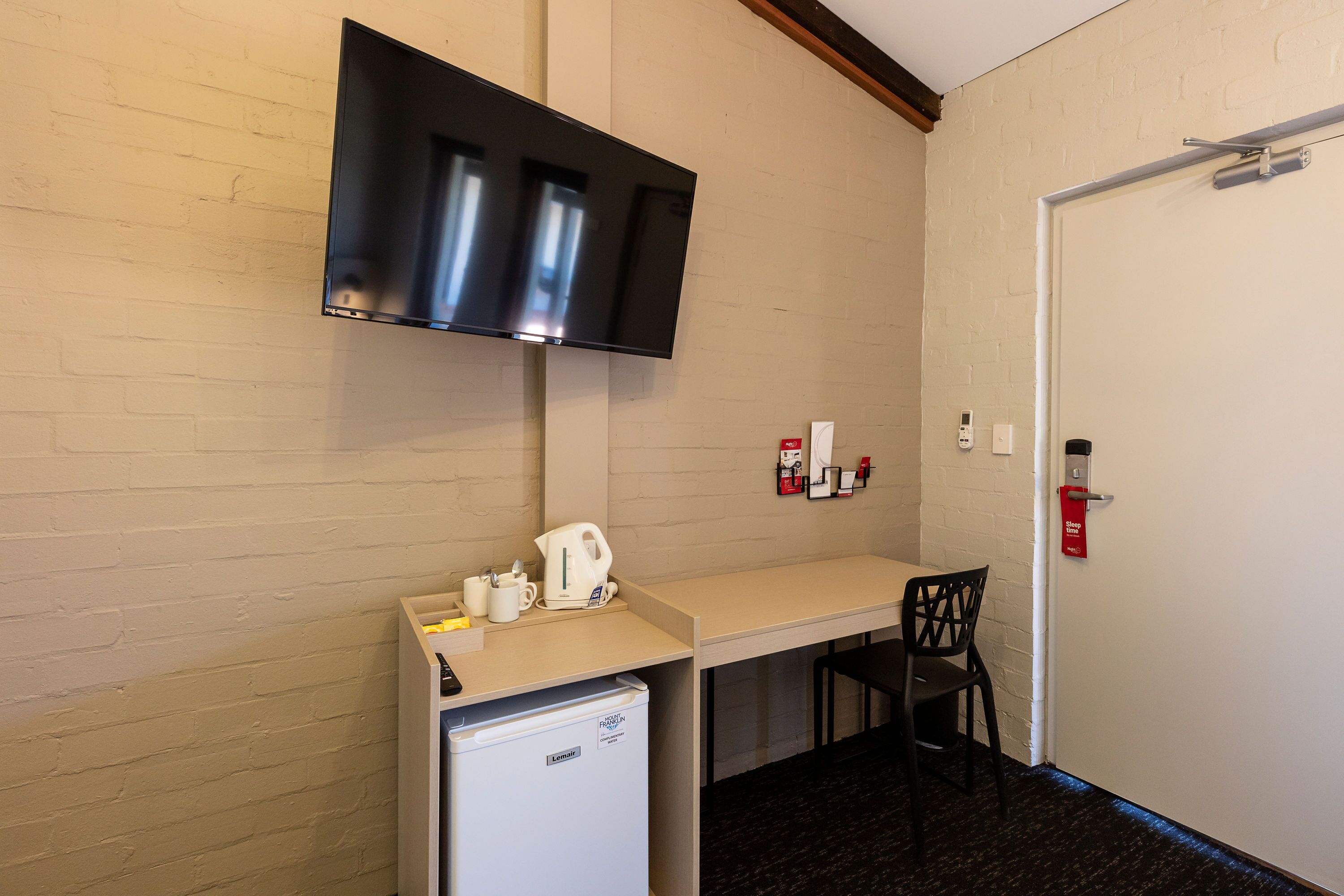 A compact hotel room with a kitchenette, a flat-screen TV mounted on the wall, a small refrigerator, a desk with a chair, and a door with a fire alarm.