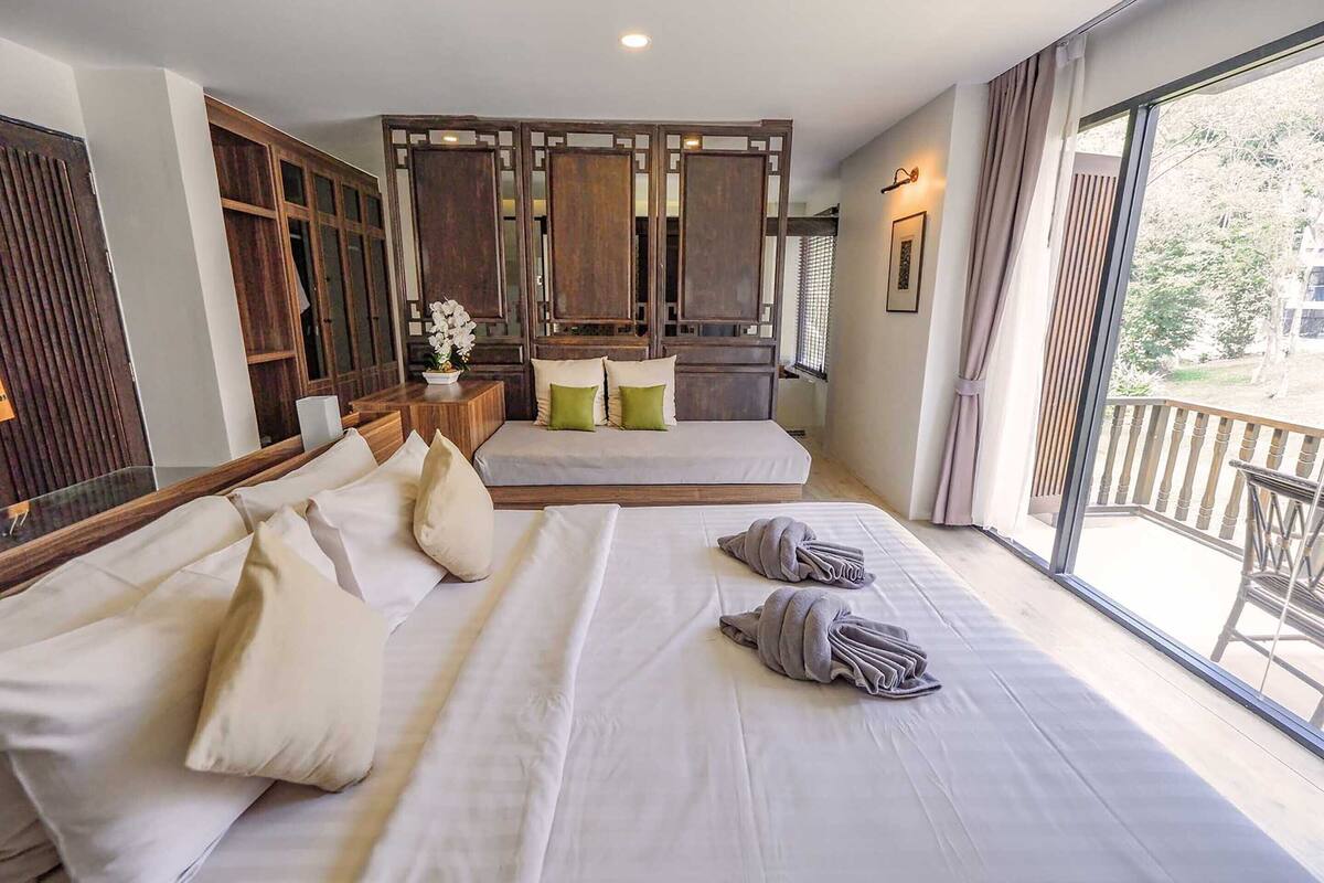 A hotel room with a large bed, a wooden partition, a balcony with a view, and a small table with a plant.