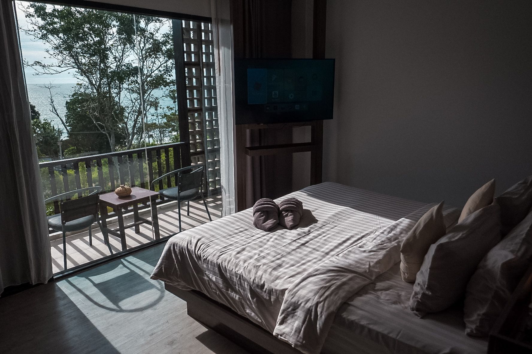 A bedroom with a bed, pillows, a TV, a balcony with a table and chairs, and a view of the sea.