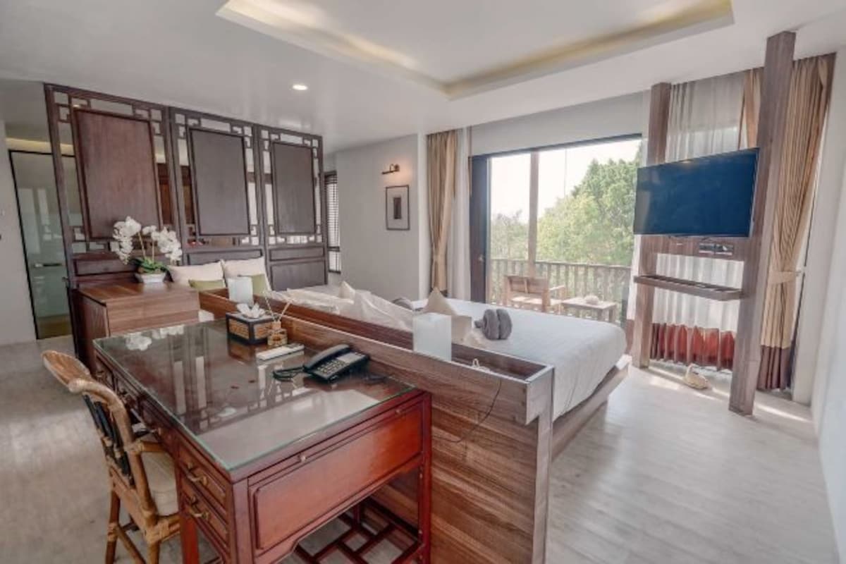 Una habitación de hotel moderna con una mesa de comedor de vidrio, muebles de madera, una cama grande, un televisor y un balcón con vistas a árboles.