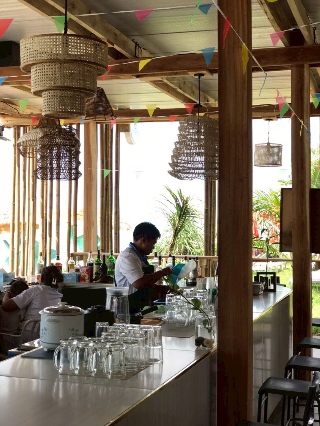 Zona de bar con lámparas de mimbre colgantes, una barra con vasos y botellas, y vistas al exterior.