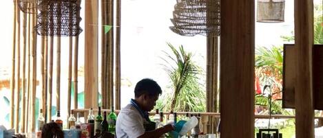 Zona de bar con lámparas de mimbre colgantes, una barra con vasos y botellas, y vistas al exterior.