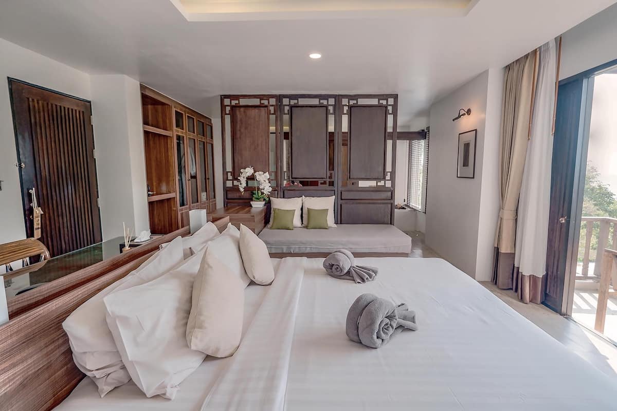 A hotel room with a large bed, a wooden wardrobe, a seating area with cushions, and a view through a window.