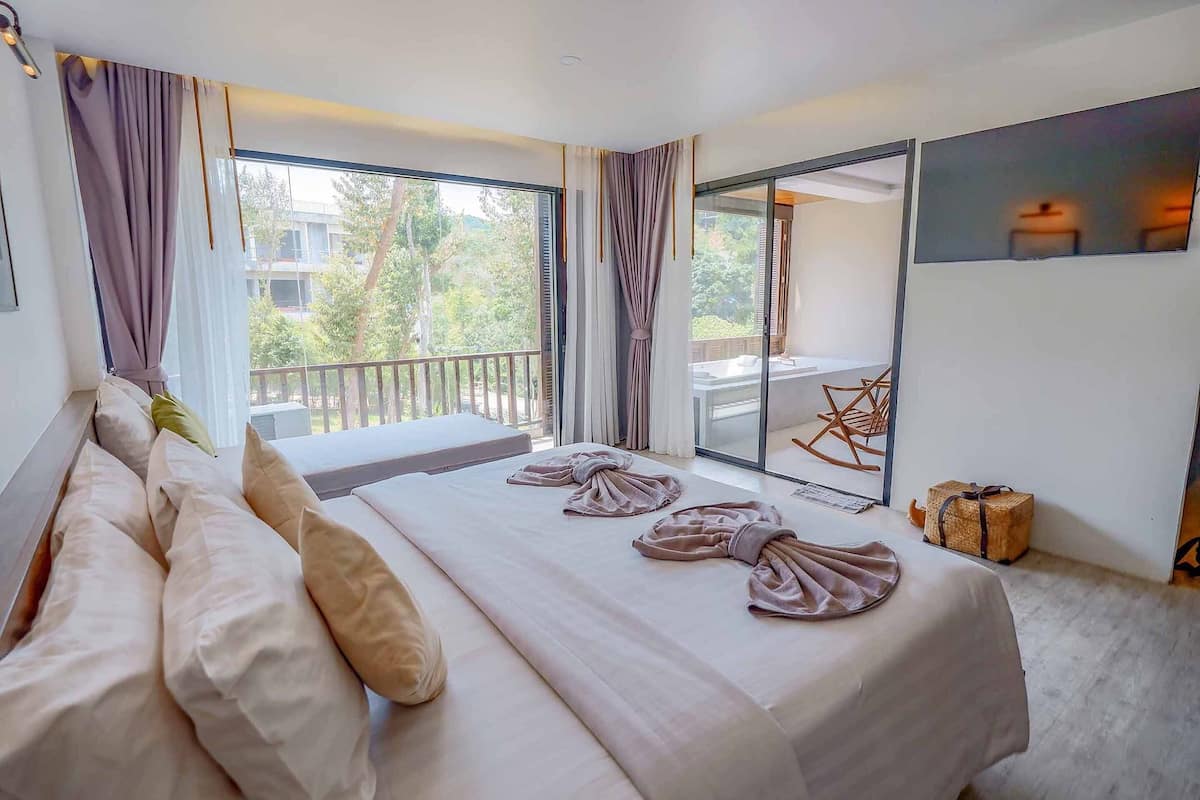 A modern hotel room with a large bed, a balcony with a view of greenery, and a sliding glass door leading to an outdoor area.