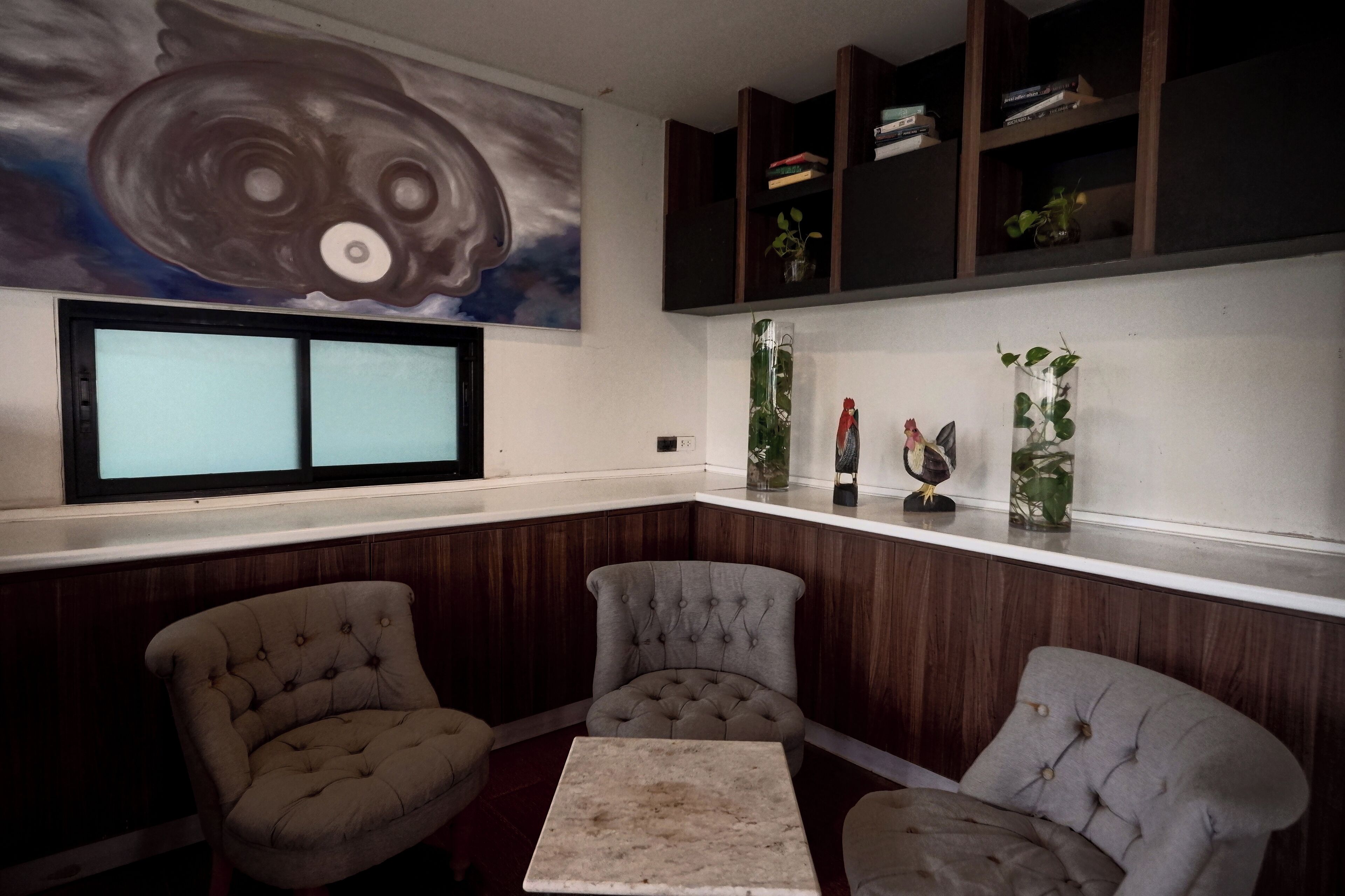 Un coin salon avec deux fauteuils et une petite table, un grand tableau au mur et une étagère avec des livres et des plantes.