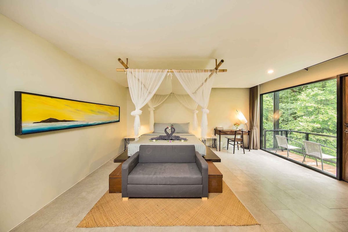 A bedroom with a bed, a gray sofa, a painting, and a balcony with chairs.