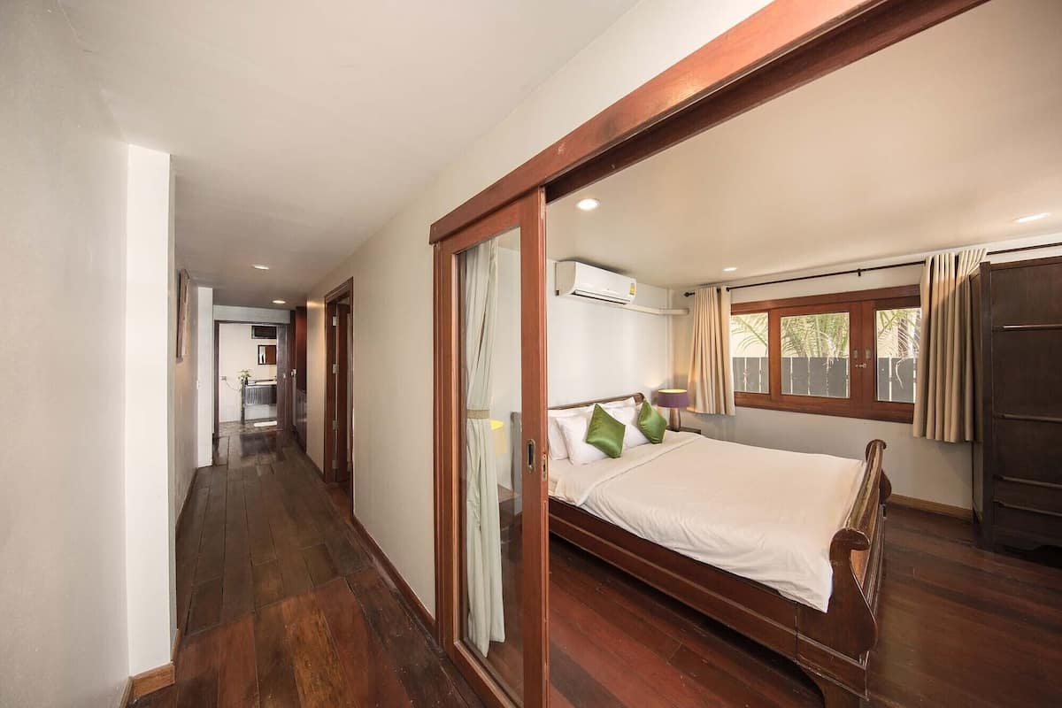 A hotel room with a wooden bed, a window with curtains, a door, and a hallway leading to another room.
