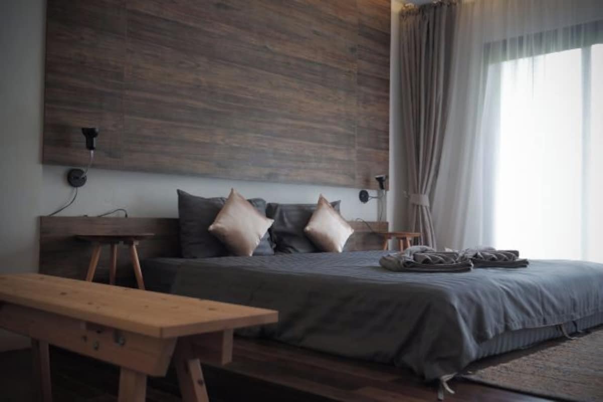 Un dormitorio con cabecera de madera, cama con ropa de cama gris, una banca de madera y una mesita de noche.