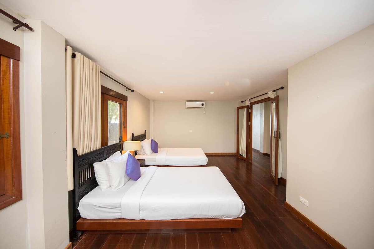 A hotel room with two beds, wooden flooring, a window with curtains, an air conditioning unit, and a door leading to another room.