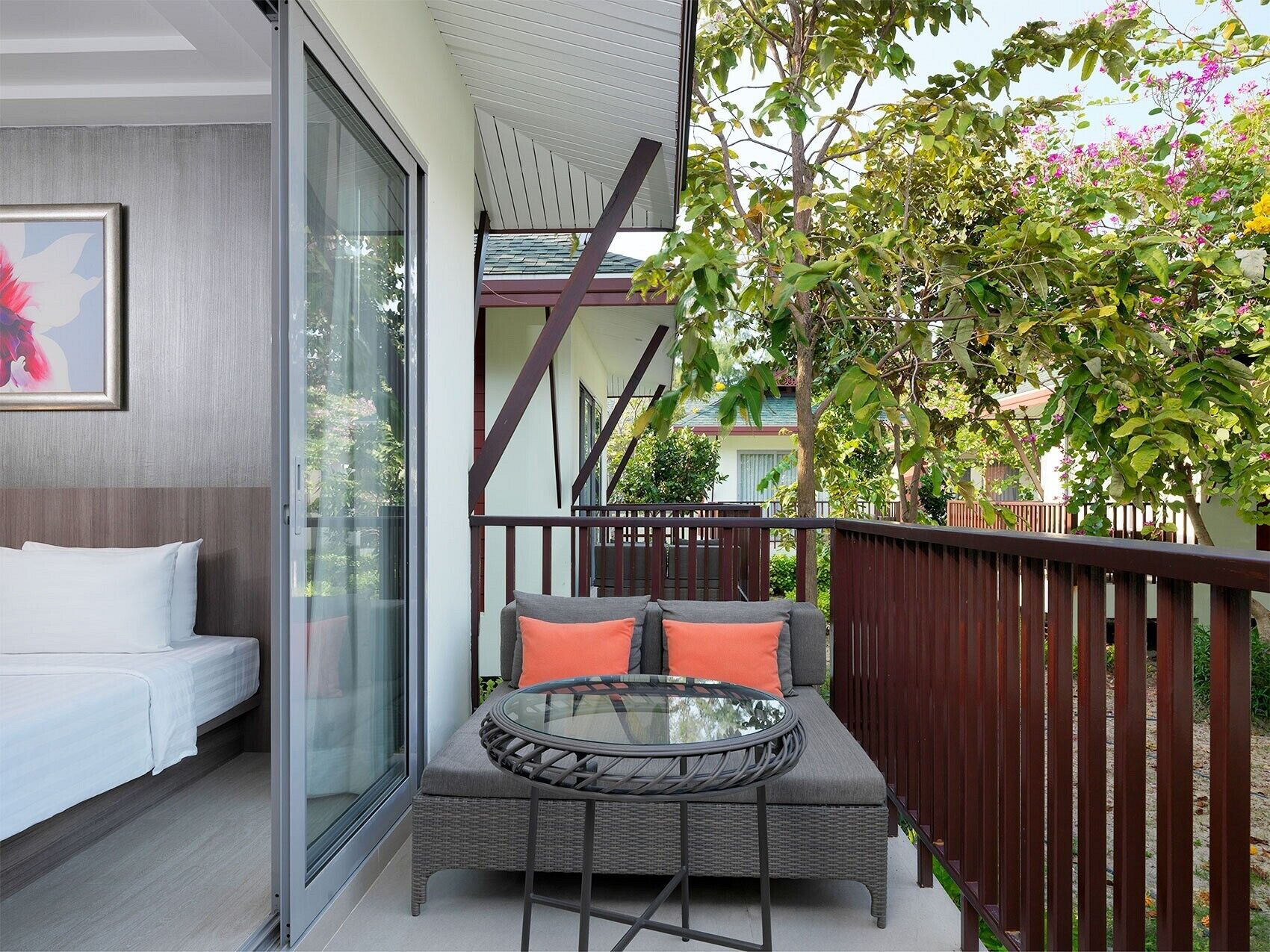 A balcony with a wicker sofa and glass table, a white bed, and a view of a neighboring building and trees.