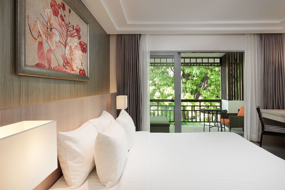 A hotel room with a large bed, a desk with a chair, a balcony with a view of trees, and a painting on the wall.
