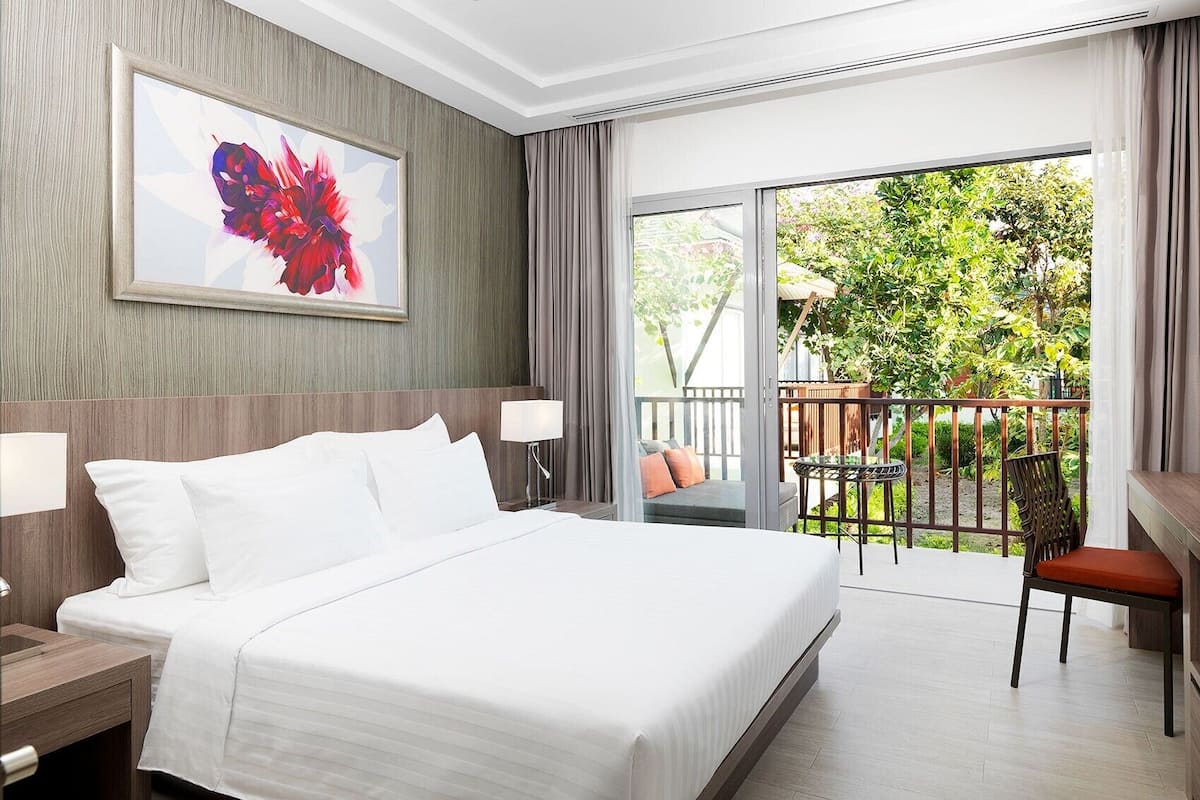 A hotel room with a large bed, a desk, a chair, a balcony with a view of greenery, and a framed artwork on the wall.