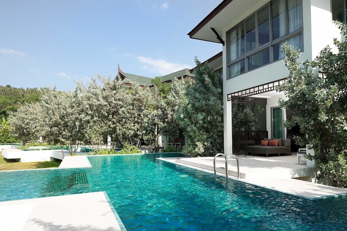 A modern pool area with clear blue water, surrounded by lush greenery and a white building with large windows.