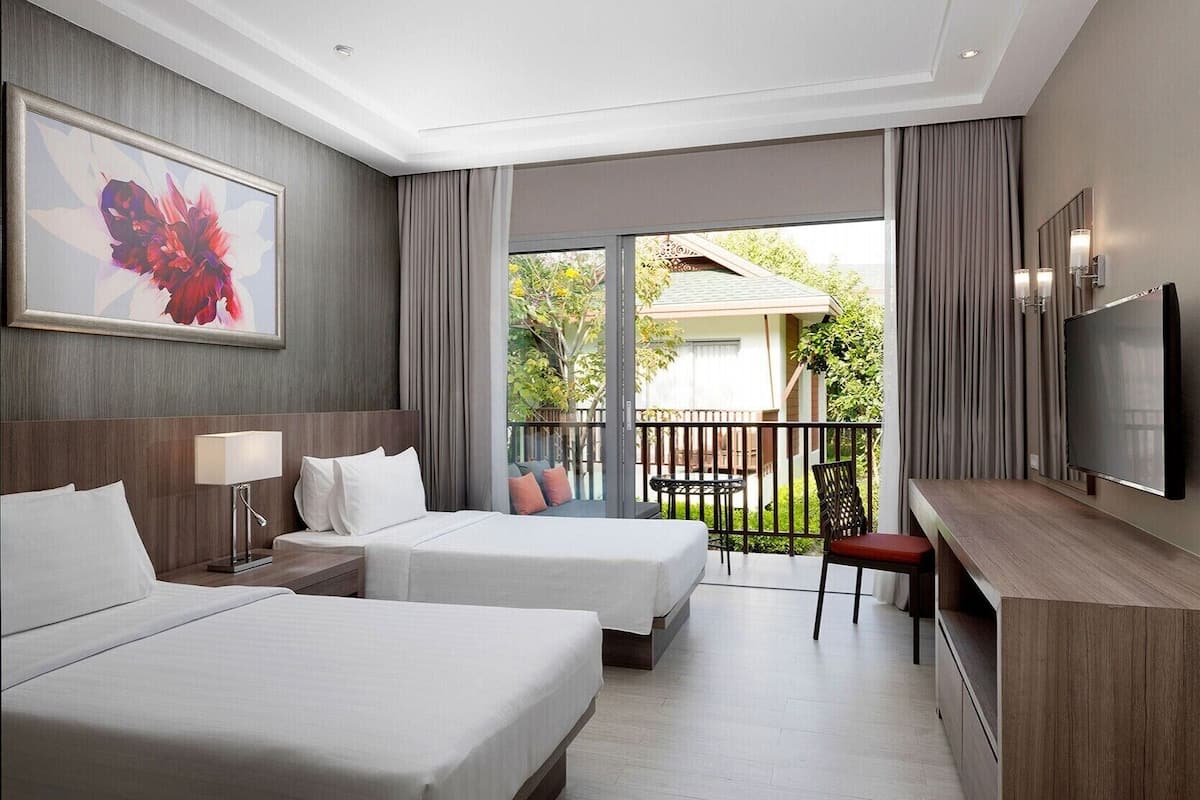 A hotel room with two beds, a TV, a desk, and a balcony with a view of greenery.