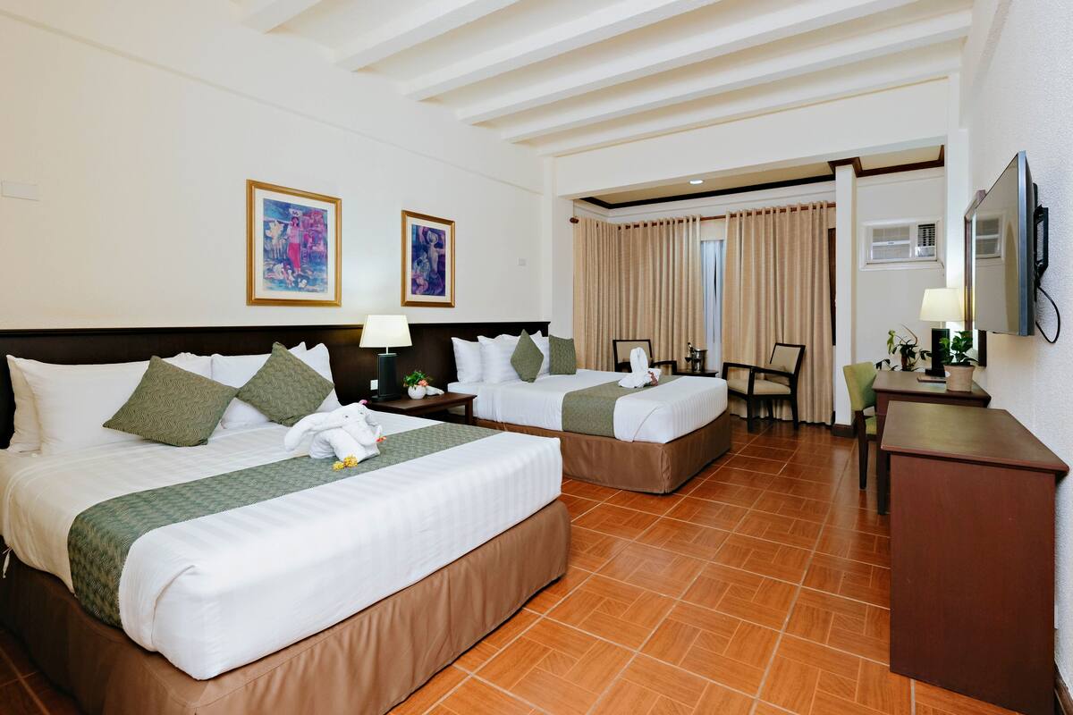 A hotel room with two beds, a desk, a chair, a TV, and artwork on the walls.