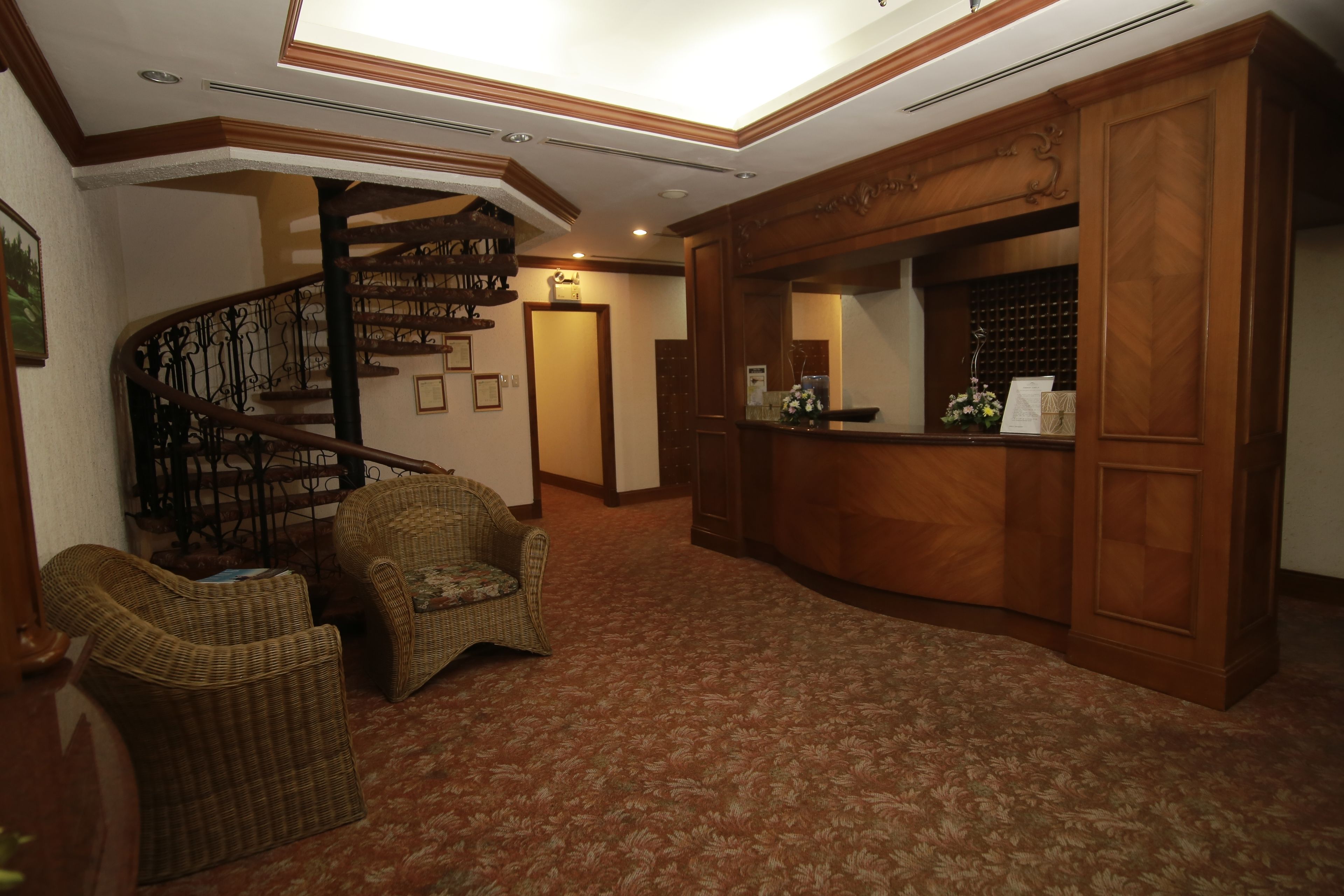 A lobby with a wooden reception desk, wicker chairs, a spiral staircase, and patterned carpet.