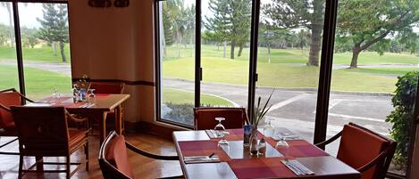 A dining area with tables and chairs, large windows offering a view of a golf course.