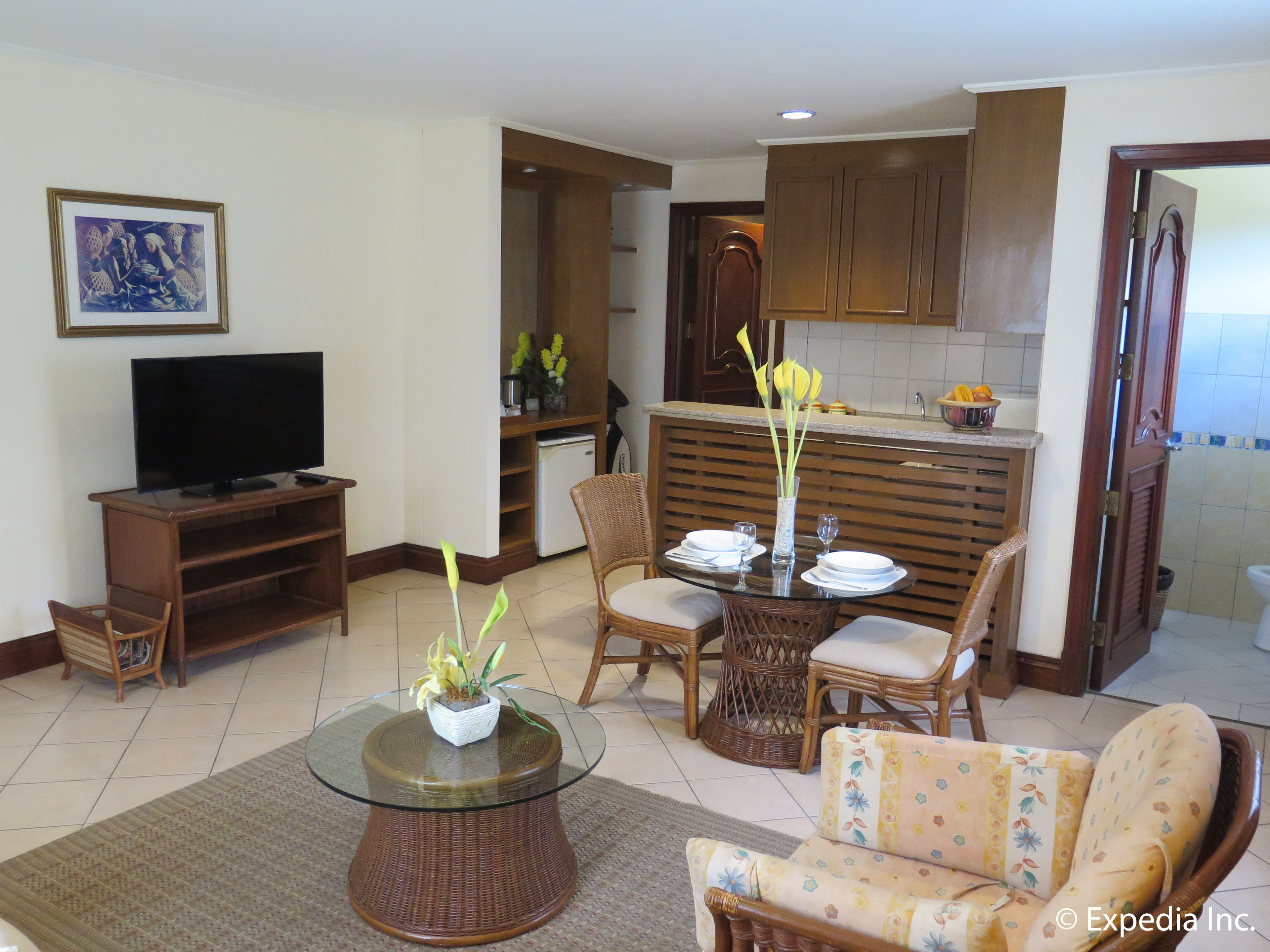 A living room with a glass coffee table, wicker furniture, a television, and a kitchen area with wooden cabinets and a refrigerator.